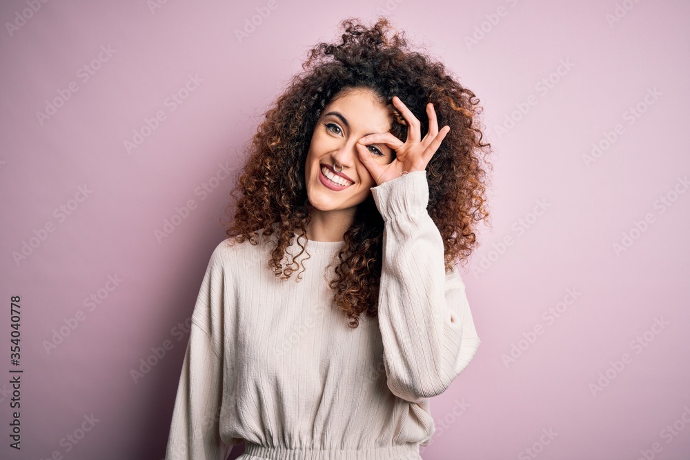 Beautiful woman with curly hair and piercing wearing casual sweater over pink background doing ok gesture with hand smiling, eye looking through fingers with happy face.