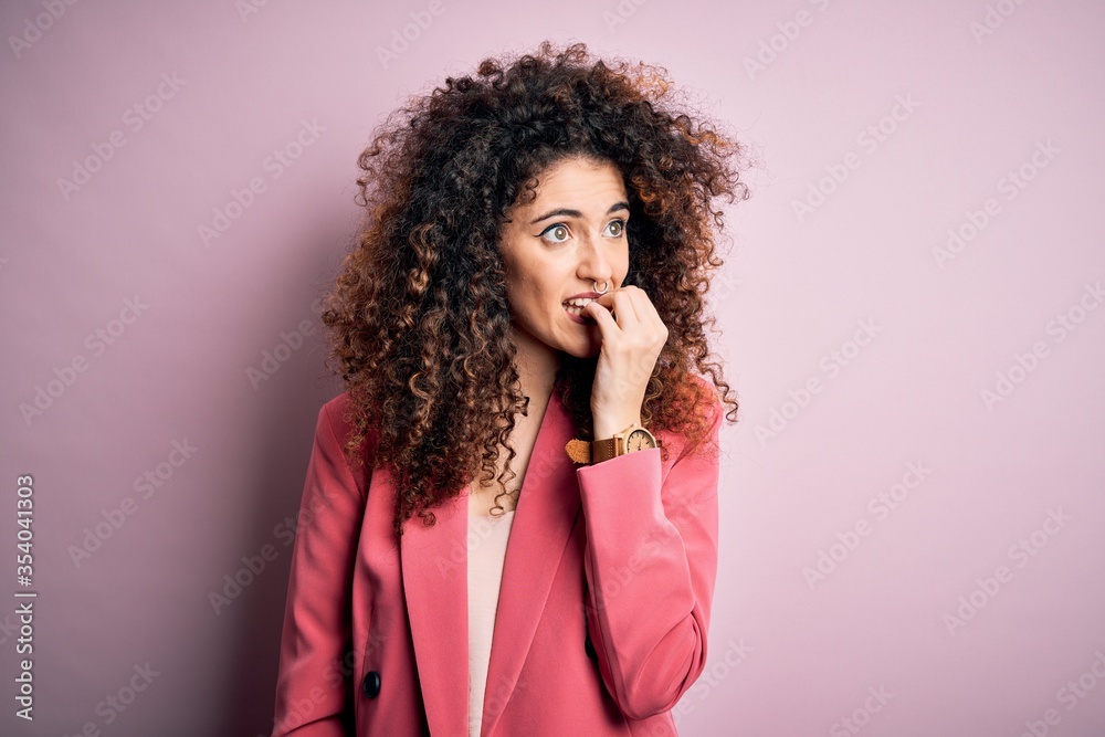 Young beautiful businesswoman with curly hair and piercing wearing elegant jacket looking stressed and nervous with hands on mouth biting nails. Anxiety problem. Young beautiful businesswoman with curly hair and piercing wearing elegant jacket looking stressed and nervous with hands on mouth biting nails. Anxiety problem.