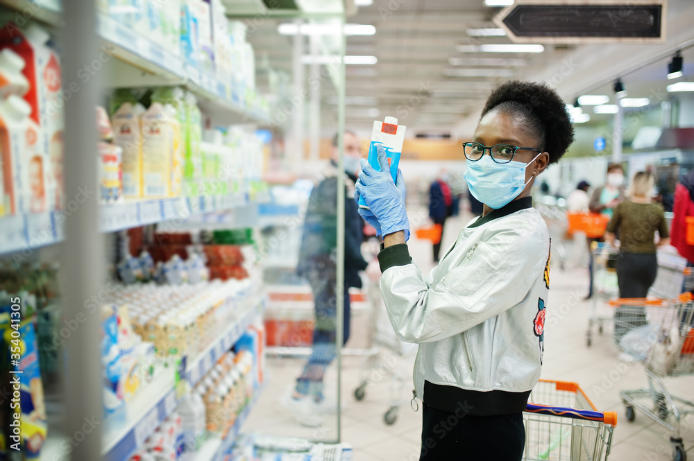 African woman wearing disposable medical mask and gloves shopping in supermarket during coronavirus pandemia outbreak. Black female choose milk at epidemic time.