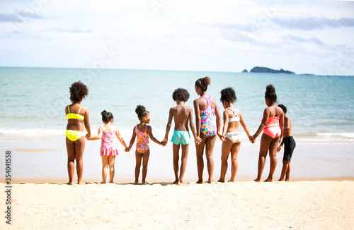 A group of children holding hands in a row on the beach in summer, rear view against sea and blue sky