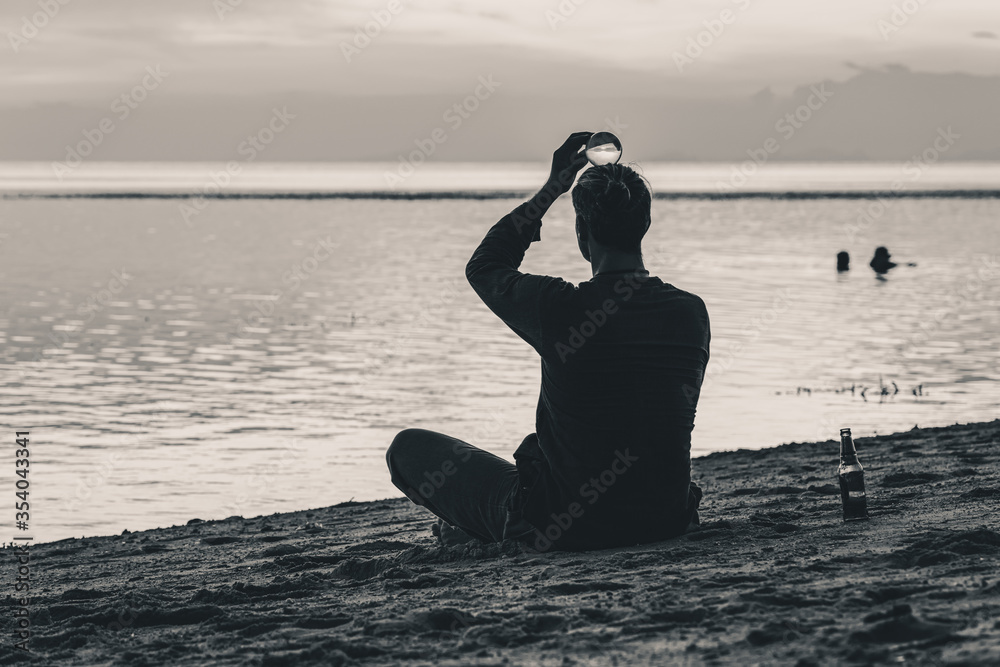 A man sits and tries to maintain balance with a transparent ball on his head on the beach at sunset