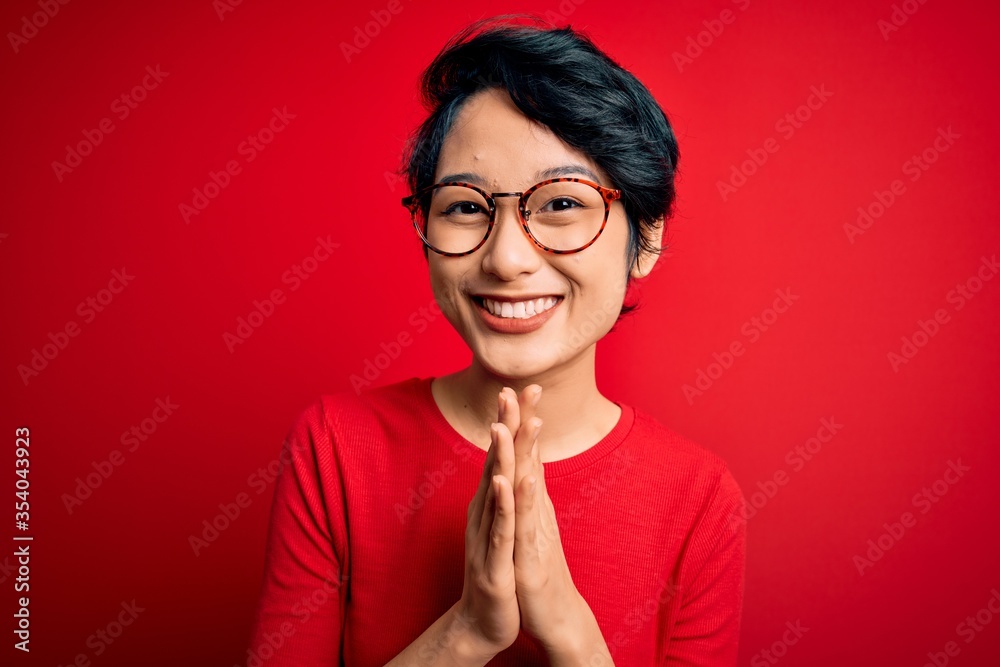 Young beautiful asian girl wearing casual t-shirt and glasses over isolated red background praying with hands together asking for forgiveness smiling confident. Young beautiful asian girl wearing casual t-shirt and glasses over isolated red background praying with hands together asking for forgiveness smiling confident.