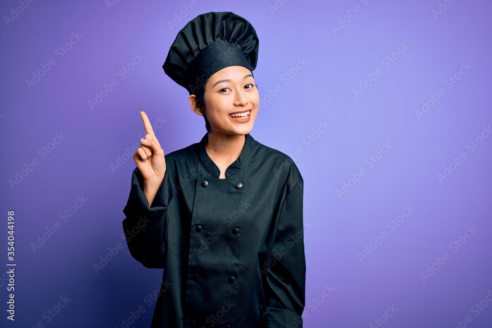 Young beautiful chinese chef woman wearing cooker uniform and hat over purple background pointing finger up with successful idea. Exited and happy. Number one. Young beautiful chinese chef woman wearing cooker uniform and hat over purple background pointing finger up with successful idea. Exited and happy. Number one.