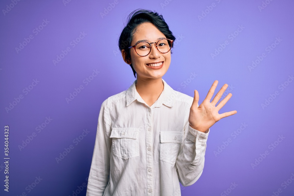 Young beautiful asian girl wearing casual shirt and glasses standing over purple background showing and pointing up with fingers number five while smiling confident and happy. Young beautiful asian girl wearing casual shirt and glasses standing over purple background showing and pointing up with fingers number five while smiling confident and happy.