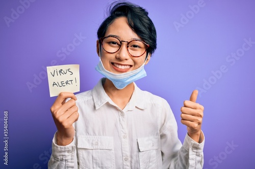 Young beautiful asian girl wearing medical mask holding reminder with virus alert message happy with big smile doing ok sign  thumb up with fingers  excellent sign