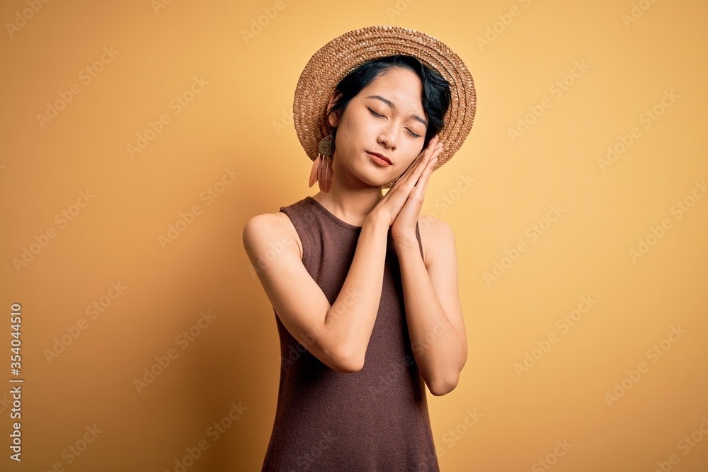 Young beautiful asian girl wearing casual t-shirt and hat over isolated yellow background sleeping tired dreaming and posing with hands together while smiling with closed eyes. Young beautiful asian girl wearing casual t-shirt and hat over isolated yellow background sleeping tired dreaming and posing with hands together while smiling with closed eyes.