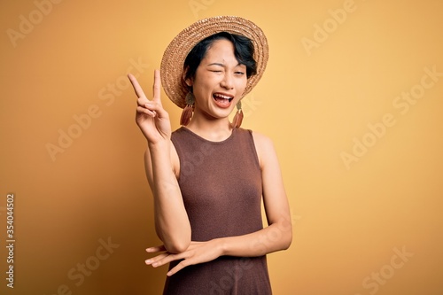 Young beautiful asian girl wearing casual t-shirt and hat over isolated yellow background smiling with happy face winking at the camera doing victory sign. Number two.