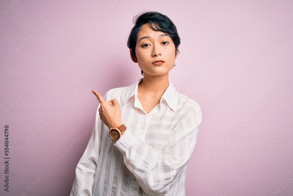 Young beautiful asian girl wearing casual shirt standing over isolated pink background Pointing with hand finger to the side showing advertisement, serious and calm face Young beautiful asian girl wearing casual shirt standing over isolated pink background Pointing with hand finger to the side showing advertisement, serious and calm face