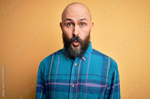 Handsome bald man with beard wearing casual shirt standing over isolated yellow background afraid and shocked with surprise expression, fear and excited face.