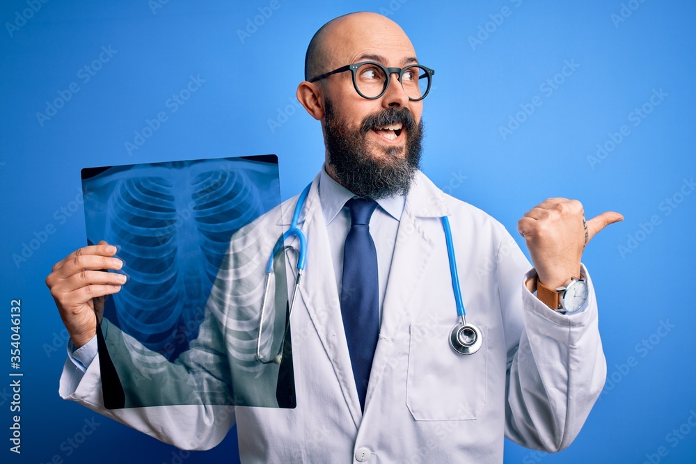 Handsome bald doctor man with beard wearing stethoscope holding chest xray pointing and showing with thumb up to the side with happy face smiling