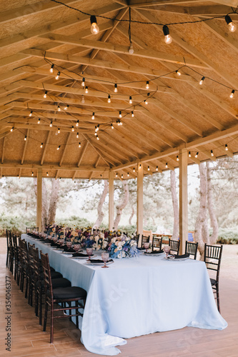 Outdoor banquet table under the wooden roof of the gazebo. Rack and cutlery, purple glasses. Floral arrangement of violet, blue, black, silver flowers. Black banquet chairs. On the table is a blue tab
