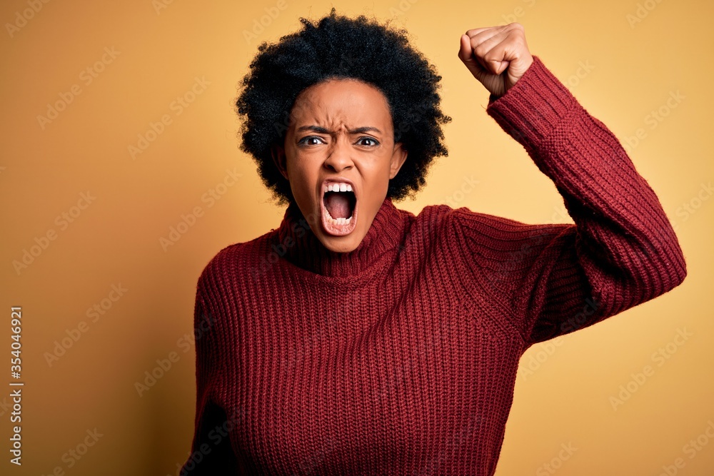 Young beautiful African American afro woman with curly hair wearing casual turtleneck sweater angry and mad raising fist frustrated and furious while shouting with anger. Rage and aggressive concept. Young beautiful African American afro woman with curly hair wearing casual turtleneck sweater angry and mad raising fist frustrated and furious while shouting with anger. Rage and aggressive concept.