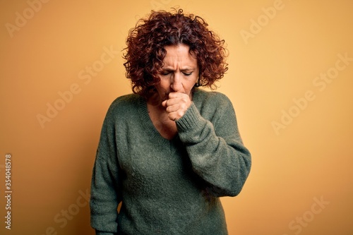 Middle age beautiful curly hair woman wearing casual sweater over isolated yellow background feeling unwell and coughing as symptom for cold or bronchitis. Health care concept.