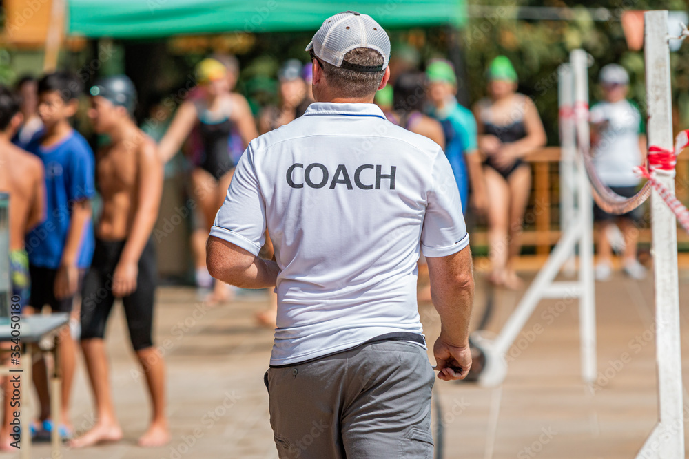 Back of male swimming coach walking on the poolside toward his young swimmers Back of male swimming coach walking on the poolside toward his young swimmers