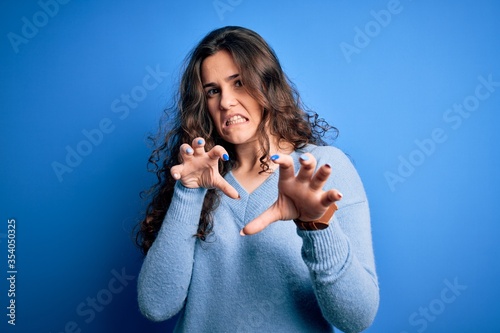 Young beautiful woman with curly hair wearing blue casual sweater over isolated background smiling funny doing claw gesture as cat, aggressive and sexy expression