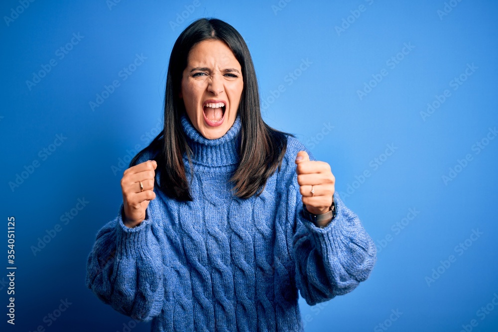 Young brunette woman with blue eyes wearing casual turtleneck sweater angry and mad raising fists frustrated and furious while shouting with anger. Rage and aggressive concept. Young brunette woman with blue eyes wearing casual turtleneck sweater angry and mad raising fists frustrated and furious while shouting with anger. Rage and aggressive concept.