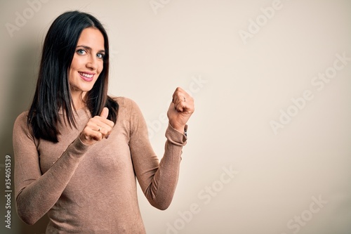 Young brunette woman with blue eyes wearing casual sweater over isolated white background Pointing to the back behind with hand and thumbs up, smiling confident photo