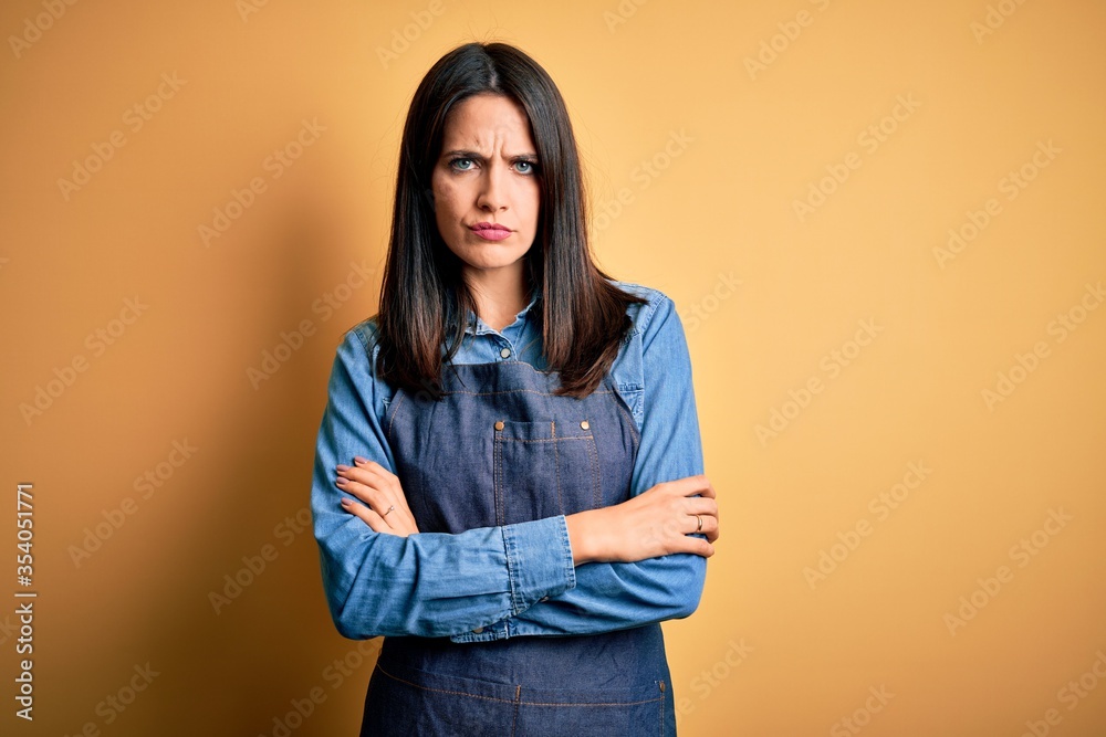 Young baker woman with blue eyes wearing apron standing over isolated yellow background skeptic and nervous, disapproving expression on face with crossed arms. Negative person. Young baker woman with blue eyes wearing apron standing over isolated yellow background skeptic and nervous, disapproving expression on face with crossed arms. Negative person.