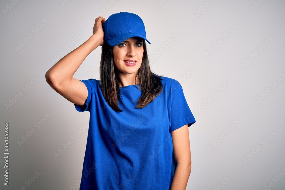 Young delivery woman with blue eyes wearing cap standing over blue background confuse and wonder about question. Uncertain with doubt, thinking with hand on head. Pensive concept.