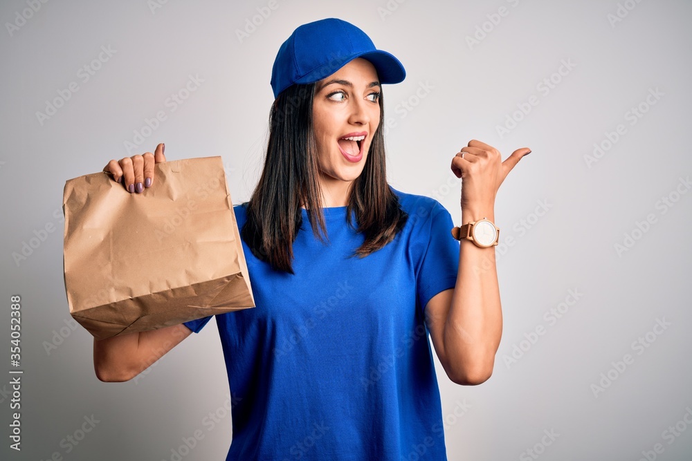 Young delivery woman with blue eyes wearing cap holding paper bag with food pointing and showing with thumb up to the side with happy face smiling Young delivery woman with blue eyes wearing cap holding paper bag with food pointing and showing with thumb up to the side with happy face smiling