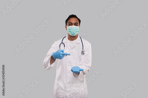 Doctor Pointing at Empty Open Hand in front of Chest Wearing Medical Mask and Gloves Isolated. Indian Man Doctor Advertisment Concept
