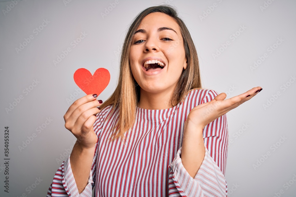 Young beautiful blonde romantic woman holding red paper heart over white background very happy and excited, winner expression celebrating victory screaming with big smile and raised hands Young beautiful blonde romantic woman holding red paper heart over white background very happy and excited, winner expression celebrating victory screaming with big smile and raised hands