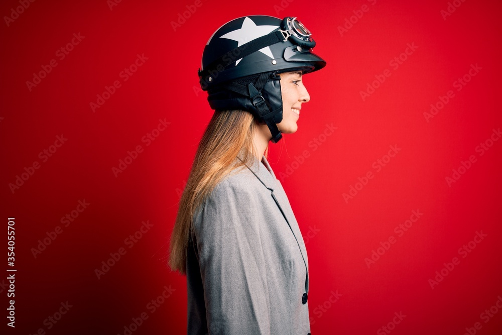 Young beautiful blonde motorcyclist woman wearing motorcycle helmet over red background looking to side, relax profile pose with natural face with confident smile.