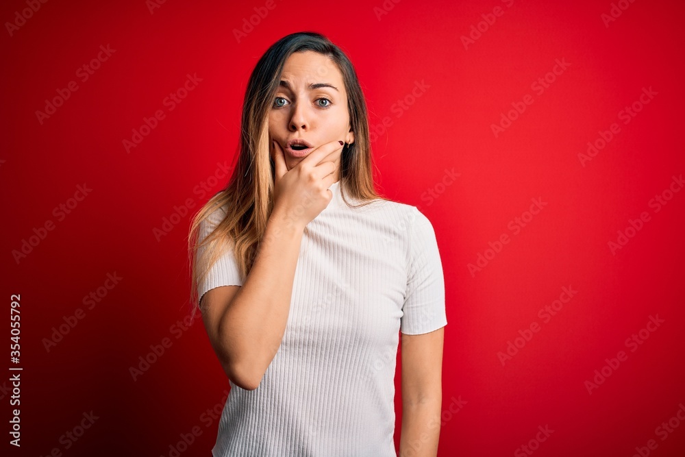 Beautiful blonde woman with blue eyes wearing casual white t-shirt over red background Looking fascinated with disbelief, surprise and amazed expression with hands on chin Beautiful blonde woman with blue eyes wearing casual white t-shirt over red background Looking fascinated with disbelief, surprise and amazed expression with hands on chin