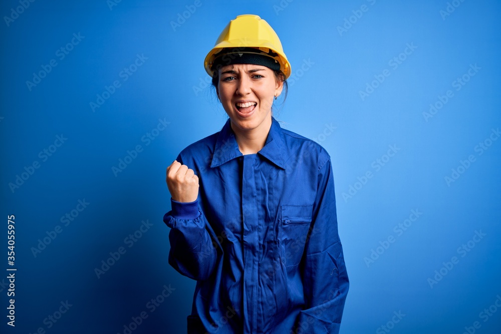Young beautiful worker woman with blue eyes wearing security helmet and uniform angry and mad raising fist frustrated and furious while shouting with anger. Rage and aggressive concept.