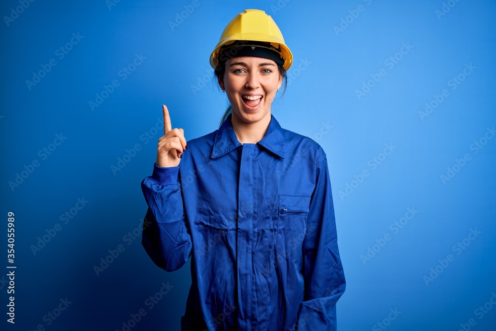 Young beautiful worker woman with blue eyes wearing security helmet and uniform pointing finger up with successful idea. Exited and happy. Number one. Young beautiful worker woman with blue eyes wearing security helmet and uniform pointing finger up with successful idea. Exited and happy. Number one.