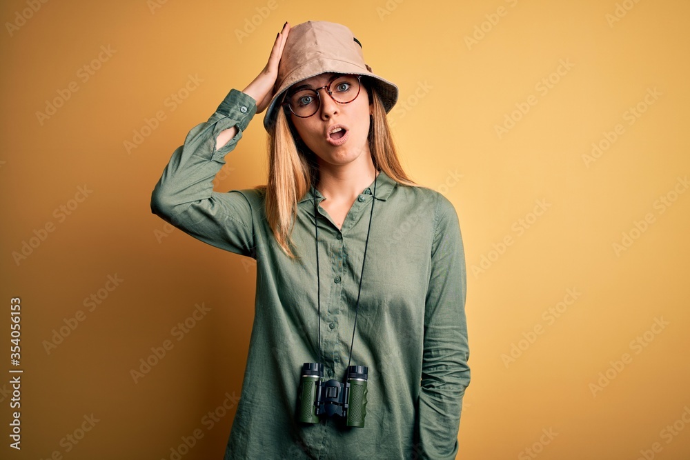 Beautiful blonde explorer woman with blue eyes wearing hat and glasses using binoculars surprised with hand on head for mistake, remember error. Forgot, bad memory concept. Beautiful blonde explorer woman with blue eyes wearing hat and glasses using binoculars surprised with hand on head for mistake, remember error. Forgot, bad memory concept.