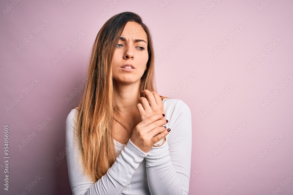 Young beautiful blonde woman with blue eyes wearing white t-shirt over pink background Suffering pain on hands and fingers, arthritis inflammation Young beautiful blonde woman with blue eyes wearing white t-shirt over pink background Suffering pain on hands and fingers, arthritis inflammation