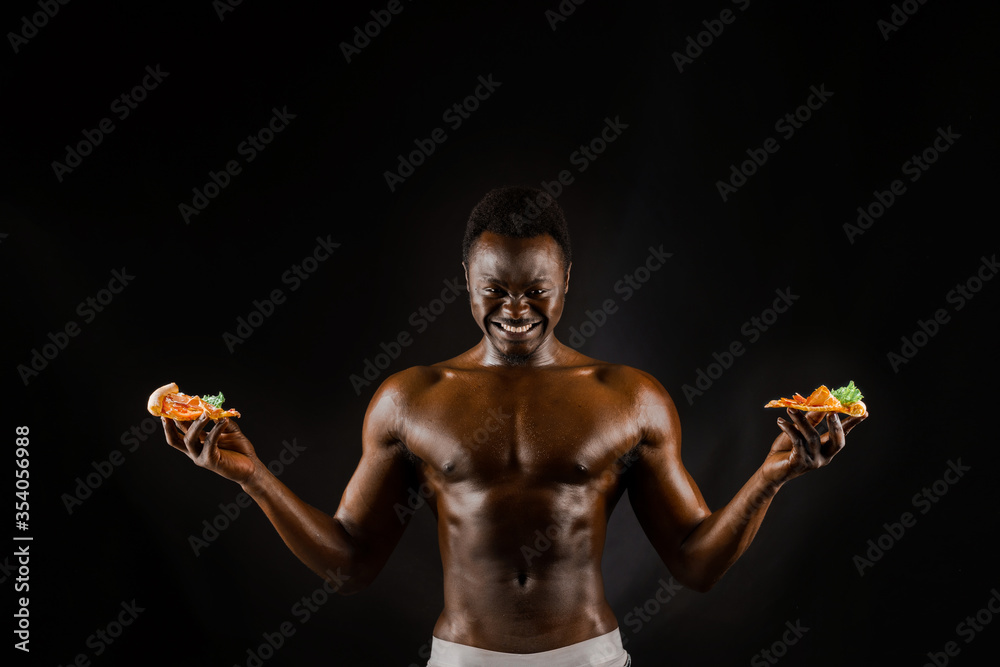Black handsome naked man with a pizza. Safety food delivery for people who stays at home. Sexy african man shows 2 pieces of pizza in his hands. Tasty food from pizzeria restaurant Black handsome naked man with a pizza. Safety food delivery for people who stays at home. Sexy african man shows 2 pieces of pizza in his hands. Tasty food from pizzeria restaurant