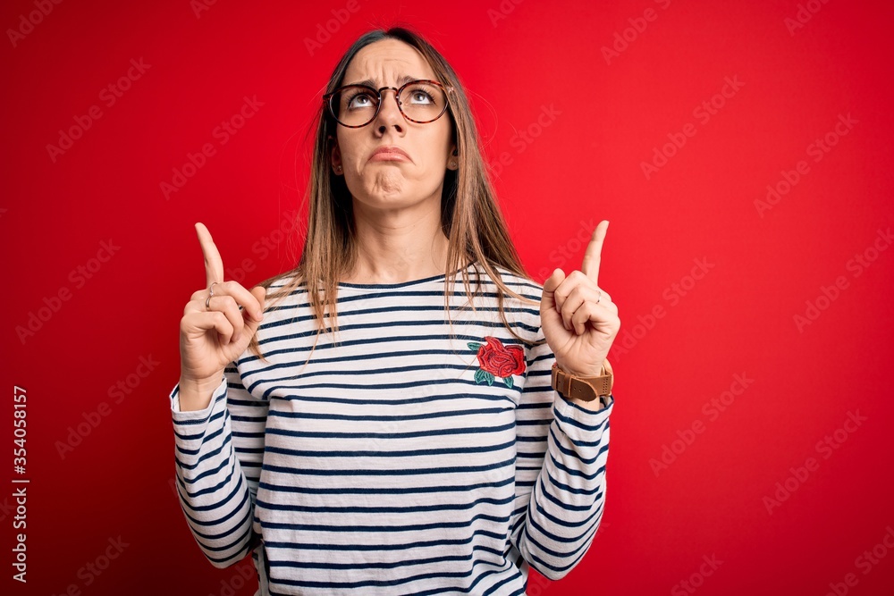 Young beautiful blonde woman with blue eyes wearing glasses standing over red background Pointing up looking sad and upset, indicating direction with fingers, unhappy and depressed. Young beautiful blonde woman with blue eyes wearing glasses standing over red background Pointing up looking sad and upset, indicating direction with fingers, unhappy and depressed.
