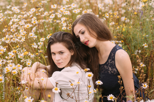 Two girls in dark blue and white dresses in sunny day sitting in chamomile field