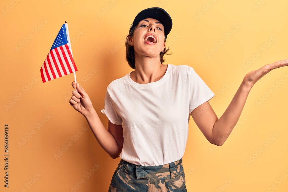 Young beautiful woman wearing usa cap holding united states flag celebrating achievement with happy smile and winner expression with raised hand