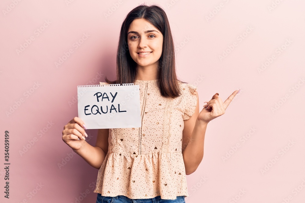 Young beautiful girl holding pay equal paper smiling happy pointing with hand and finger to the side Young beautiful girl holding pay equal paper smiling happy pointing with hand and finger to the side
