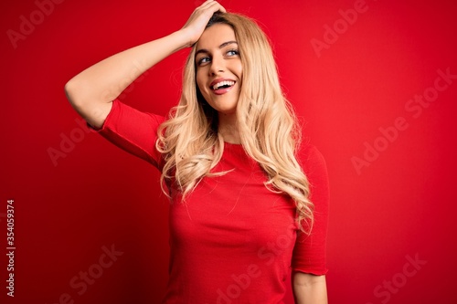 Young beautiful blonde woman wearing casual t-shirt standing over isolated red background smiling confident touching hair with hand up gesture, posing attractive and fashionable