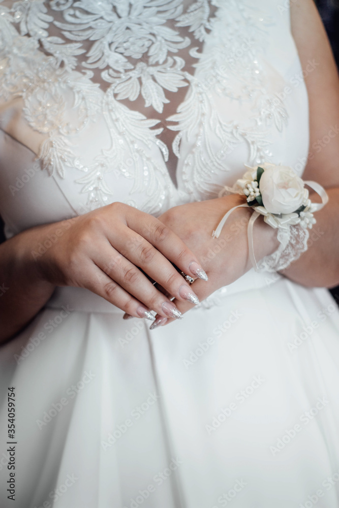 Bride holds her hands with bracelet with white flower on the belt of dress. Bride crossed her hands over the dress. Bride in a lovely white dress. Bride holds her hands with bracelet with white flower on the belt of dress. Bride crossed her hands over the dress. Bride in a lovely white dress.