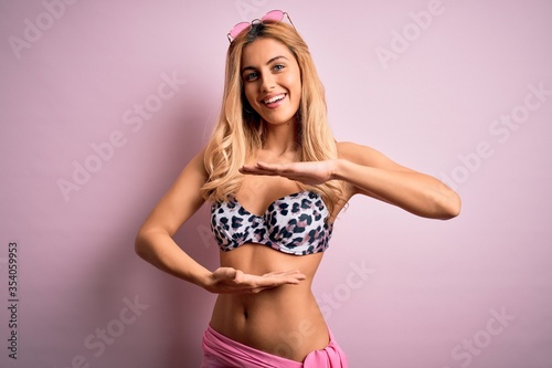 Young beautiful blonde woman on vacation wearing bikini over isolated pink background gesturing with hands showing big and large size sign, measure symbol. Smiling looking at the camera. Measuring