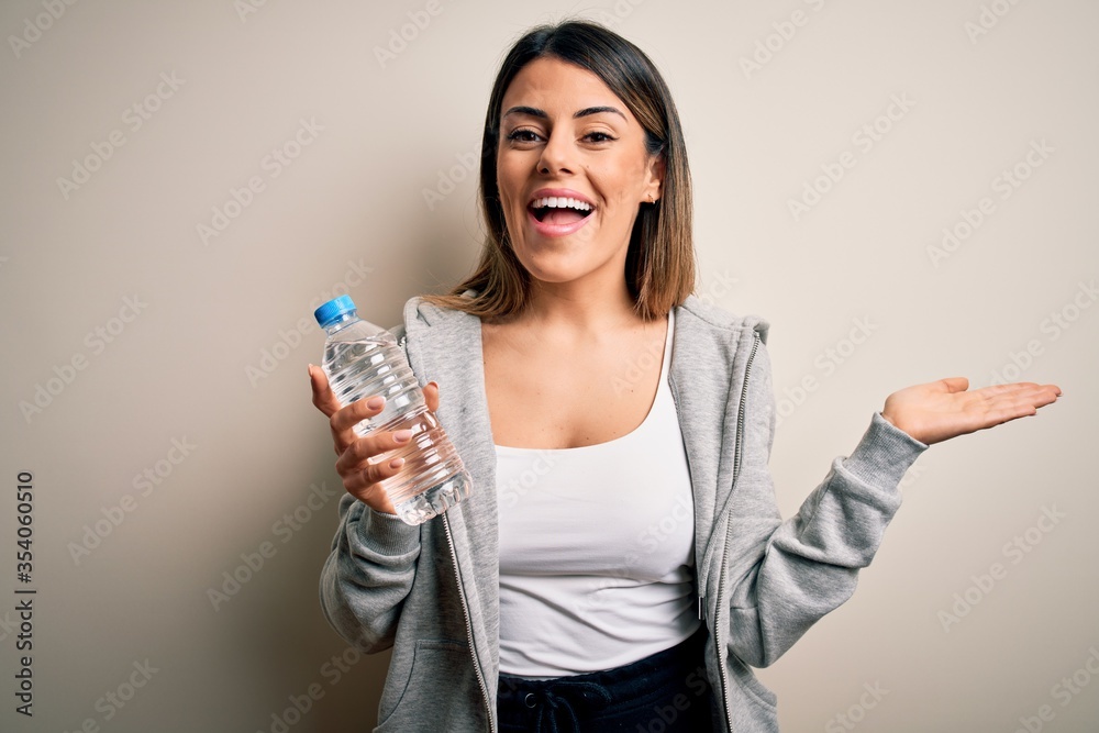 Young beautiful brunette sporty woman drinking bottle of water over isolated white background very happy and excited, winner expression celebrating victory screaming with big smile and raised hands Young beautiful brunette sporty woman drinking bottle of water over isolated white background very happy and excited, winner expression celebrating victory screaming with big smile and raised hands