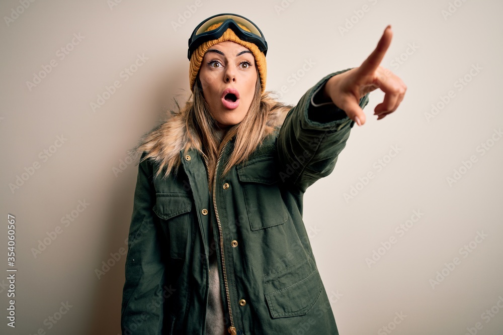 Young brunette skier woman wearing snow clothes and ski goggles over white background Pointing with finger surprised ahead, open mouth amazed expression, something on the front Young brunette skier woman wearing snow clothes and ski goggles over white background Pointing with finger surprised ahead, open mouth amazed expression, something on the front