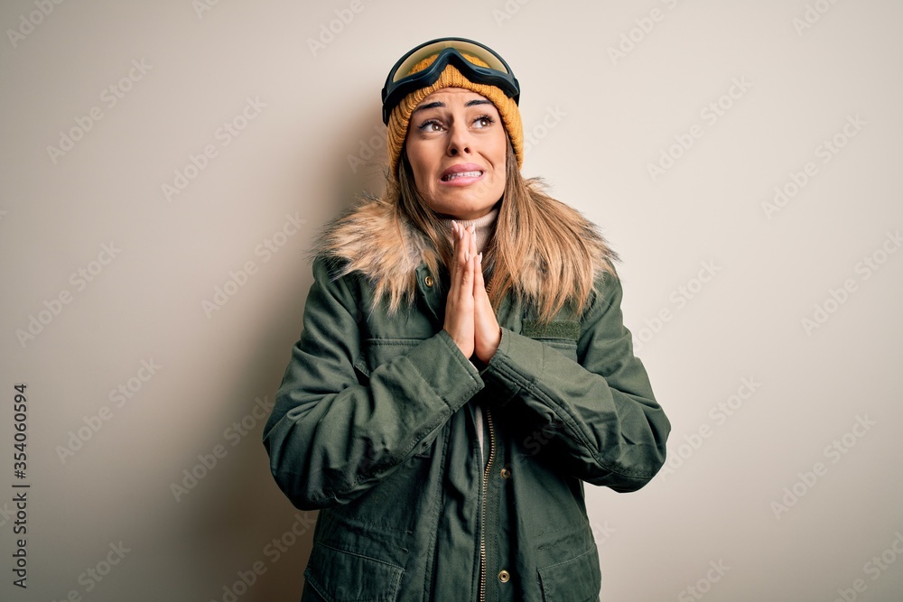 Young brunette skier woman wearing snow clothes and ski goggles over white background begging and praying with hands together with hope expression on face very emotional and worried. Begging.
