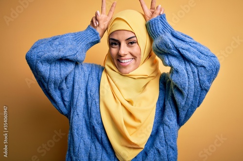 Young beautiful brunette muslim woman wearing arab hijab over isolated yellow background Posing funny and crazy with fingers on head as bunny ears, smiling cheerful