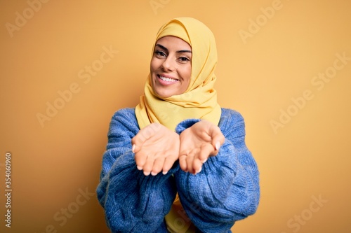 Young beautiful brunette muslim woman wearing arab hijab over isolated yellow background Smiling with hands palms together receiving or giving gesture. Hold and protection