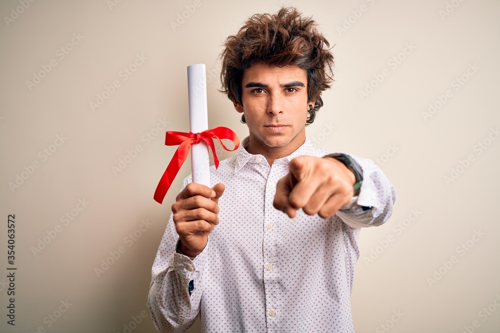 Young handsome student man holding diploma standing over isolated pink background pointing with finger to the camera and to you, hand sign, positive and confident gesture from the front Young handsome student man holding diploma standing over isolated pink background pointing with finger to the camera and to you, hand sign, positive and confident gesture from the front