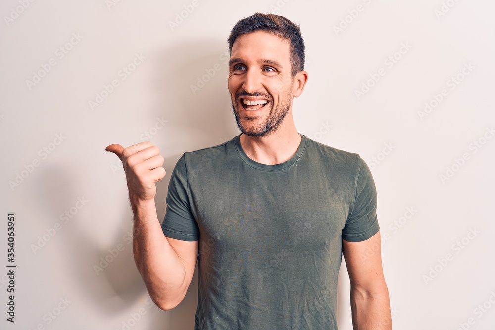Young handsome man wearing casual t-shirt standing over isolated white background pointing thumb up to the side smiling happy with open mouth