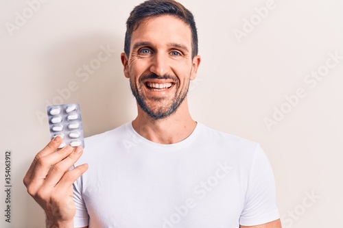Young handsome man holding medicine pills standing over isolated white background looking positive and happy standing and smiling with a confident smile showing teeth