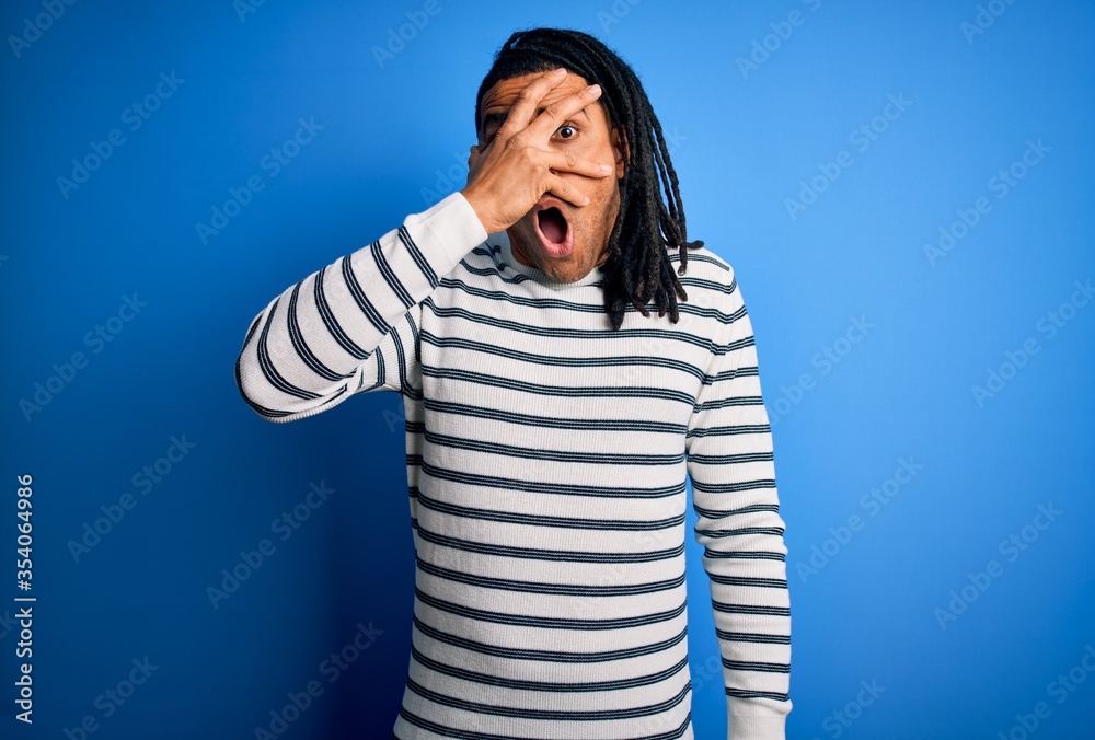 Young handsome african american afro man with dreadlocks wearing casual striped sweater peeking in shock covering face and eyes with hand, looking through fingers with embarrassed expression. Young handsome african american afro man with dreadlocks wearing casual striped sweater peeking in shock covering face and eyes with hand, looking through fingers with embarrassed expression.