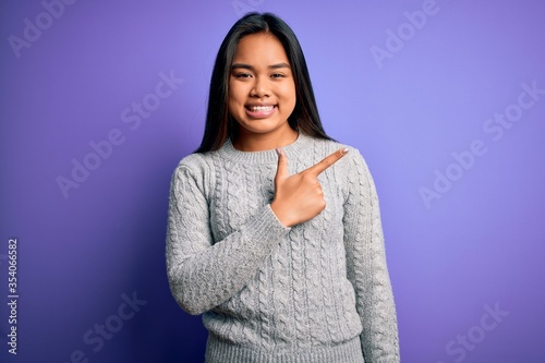 Young beautiful asian girl wearing casual sweater standing over isolated purple background cheerful with a smile of face pointing with hand and finger up to the side with happy and natural expression © Krakenimages.com